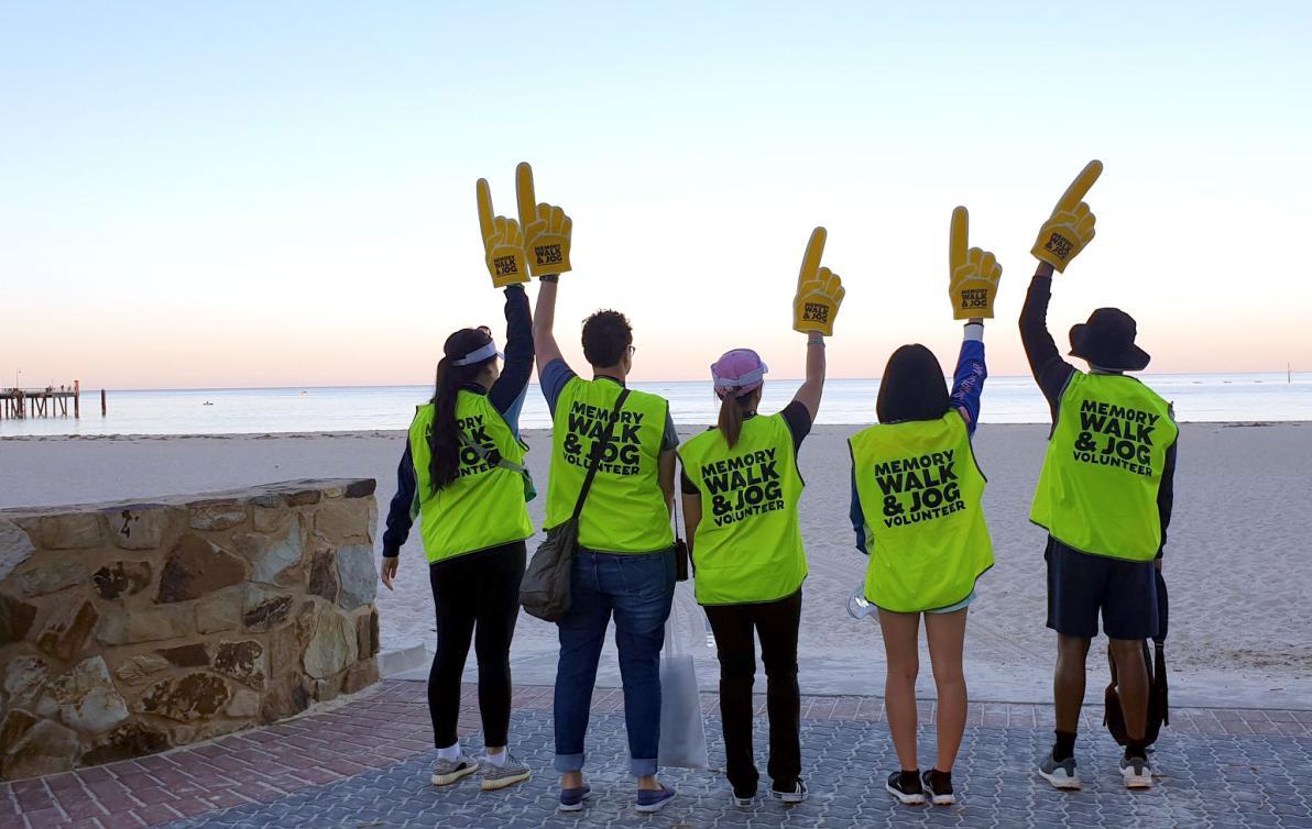 Four people with high visibility yellow vests facing away from the camera, on a beach with large foam fingers raised above their heads