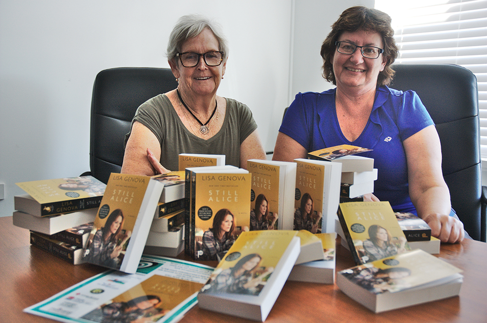 Jenny and Dot sitting in front of a pile of copies of the book Still Alice by Lisa Genova.
