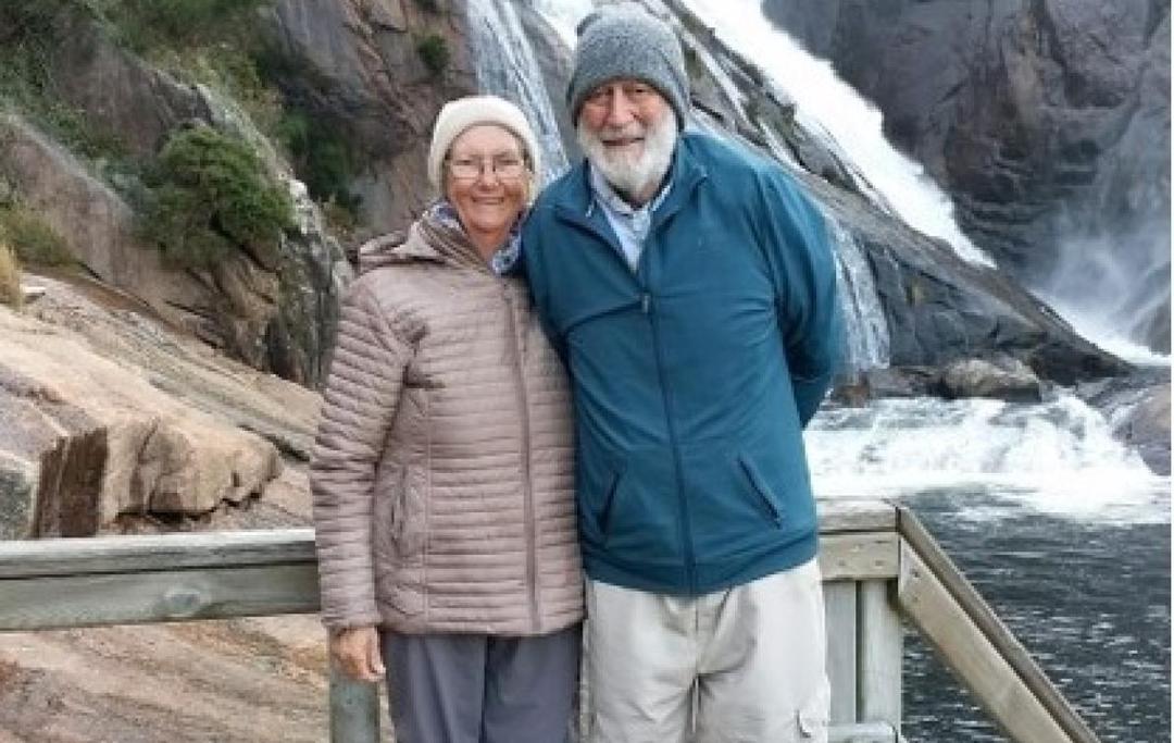 An older couple stand by a wooden fence with a waterfall in the background