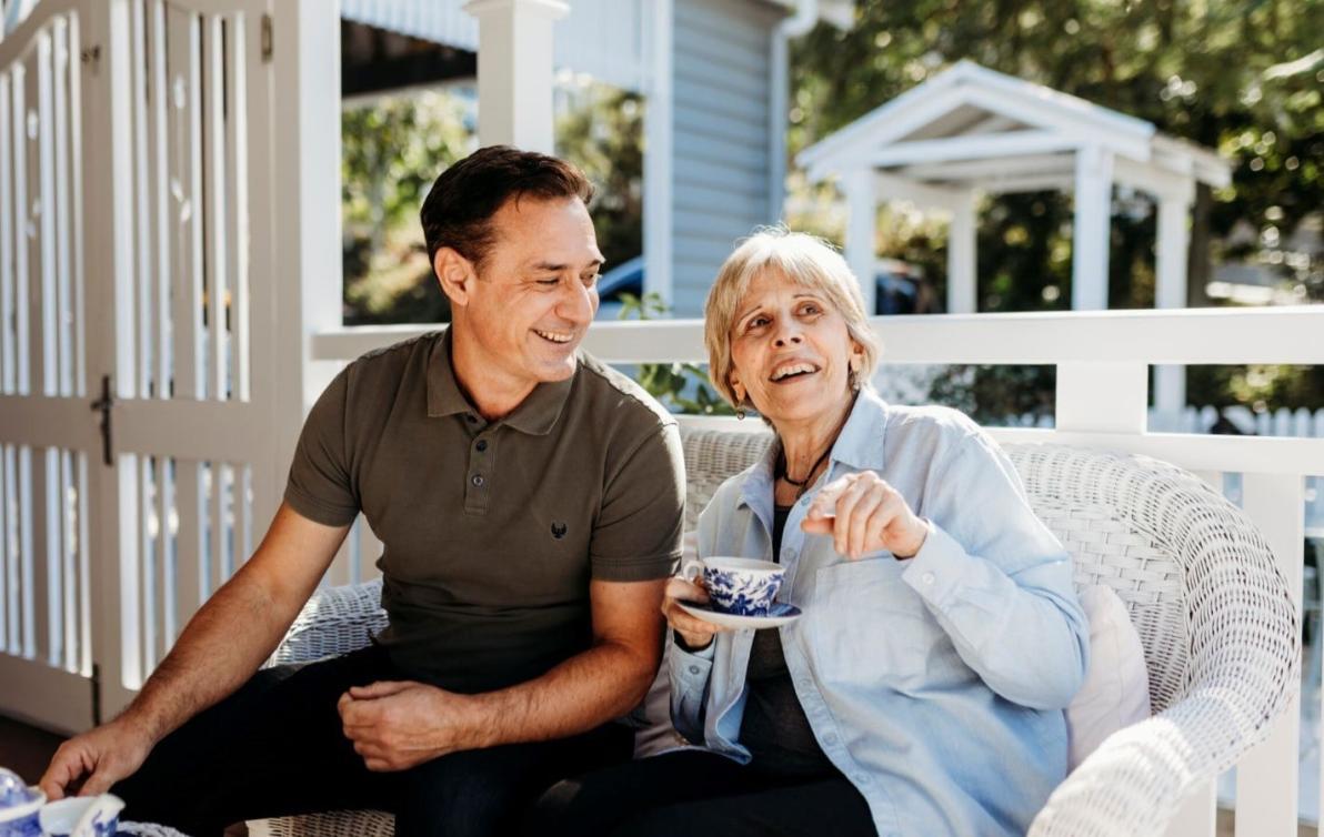 A young man and an older woman sit smiling and drinking tea on a sunny verandah.