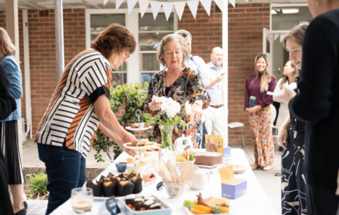 People stand around a table laid with cakes and biscuits in a sunny back yard.