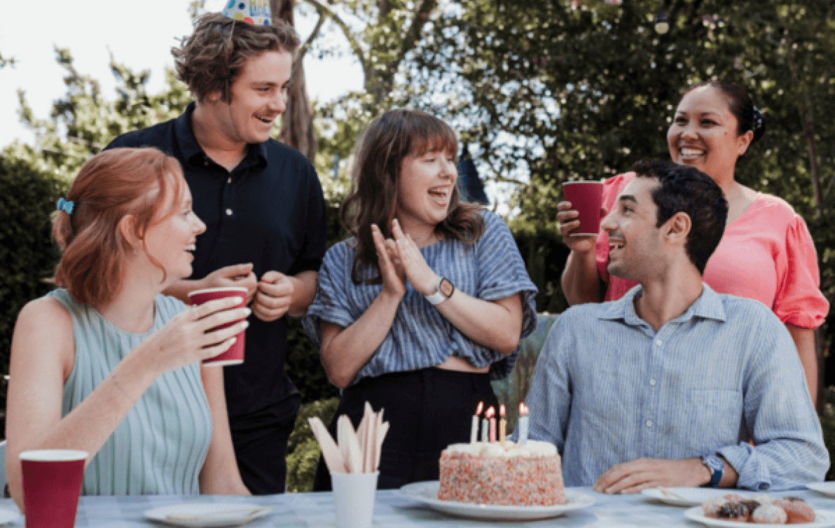 Five people sit happily around a birthday cake, with party hats and drinks.