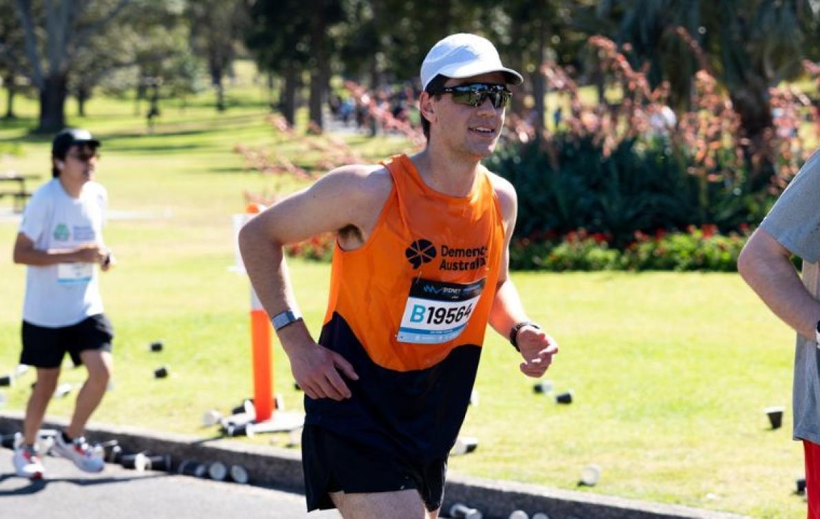 A man in Dementia Australia-branded running clothes runs through a park as others run behind him.