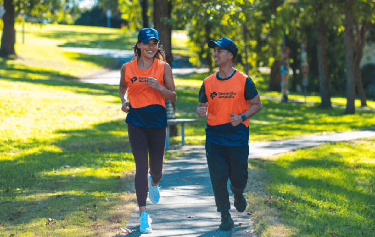 Two people in Dementia Australia-branded running clothes jog through a park, smiling.