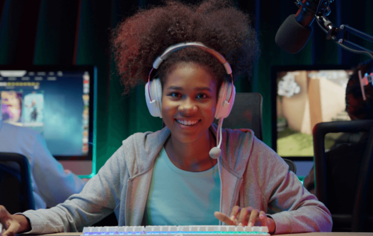 A young woman wearing gaming headphones smiles into camera as people play computer games behind her.