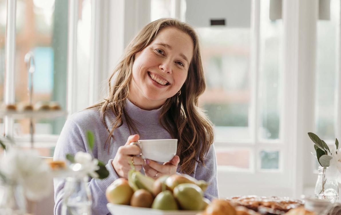 Female enjoying a cup of tea and smiling, surrounded by food on a table in a nice sunny setting at a dining table.