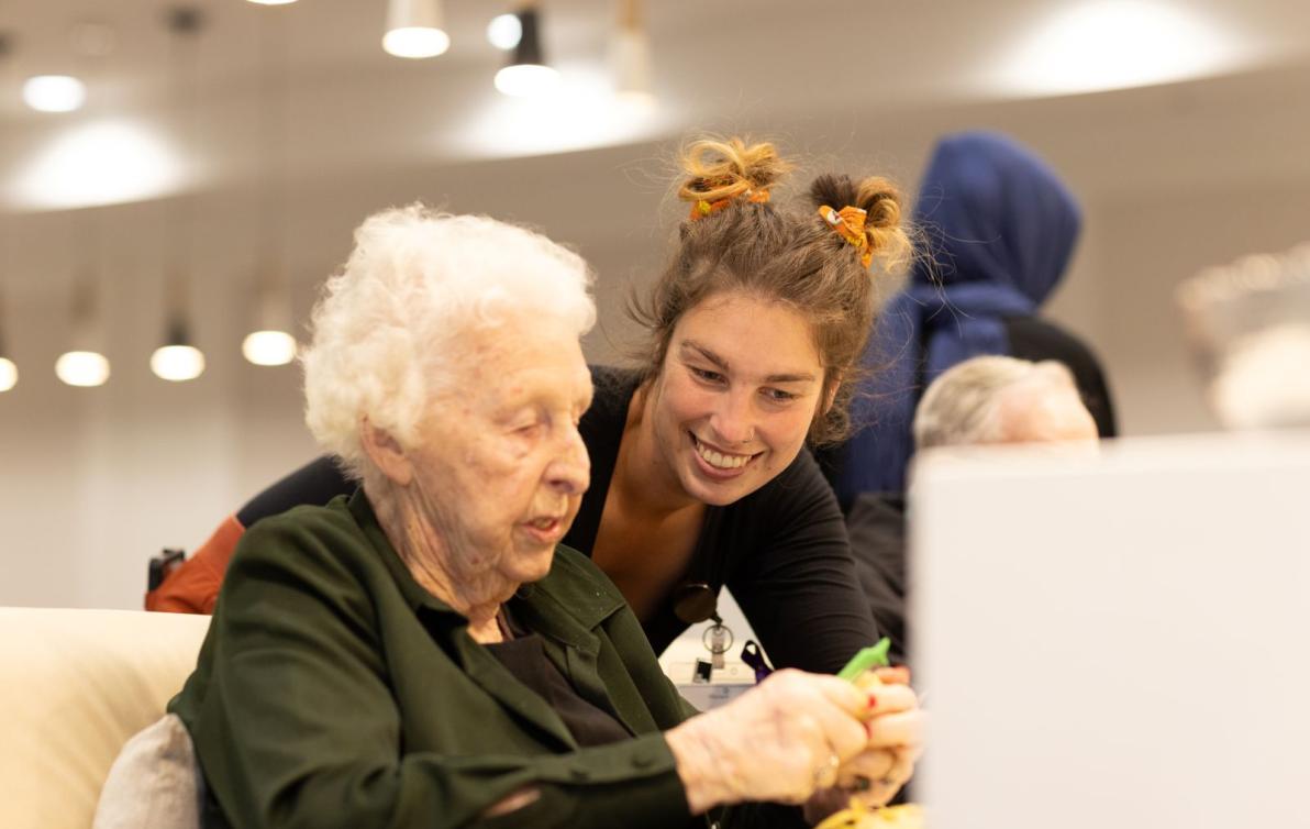 An older woman sits at a table peeling a potato while a younger woman watches over her shoulder, smiling.