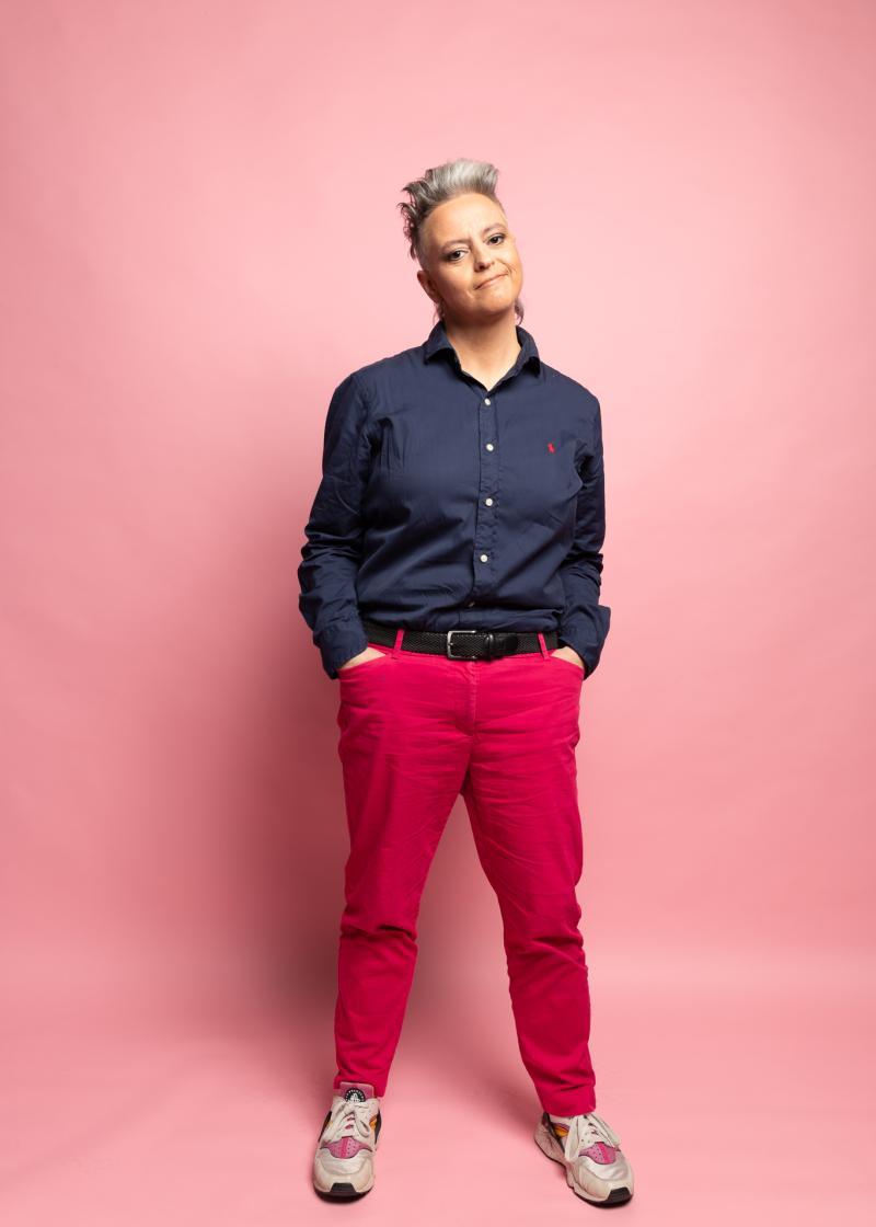 Comedian Geraldine Hickey looking at the camera wearing a dark blue shirt, red pants and sneakers, standing in front of a pink background.