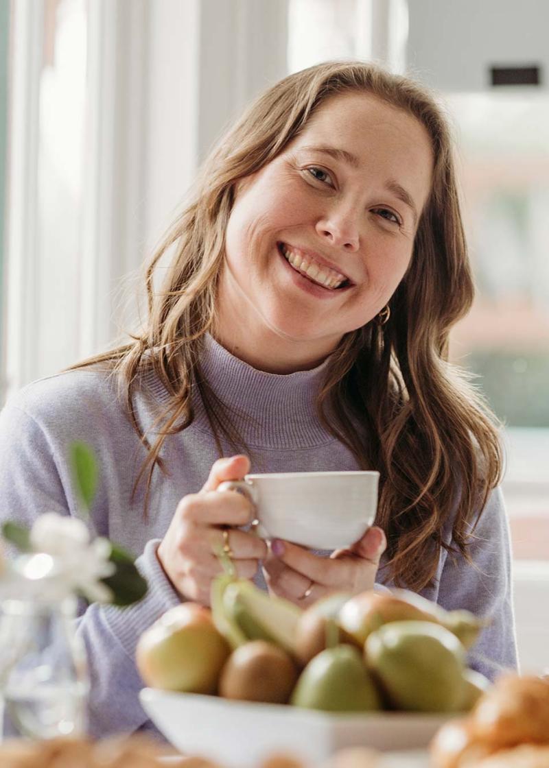 Female enjoying a cup of tea and smiling, surrounded by food on a table in a nice sunny setting at a dining table.