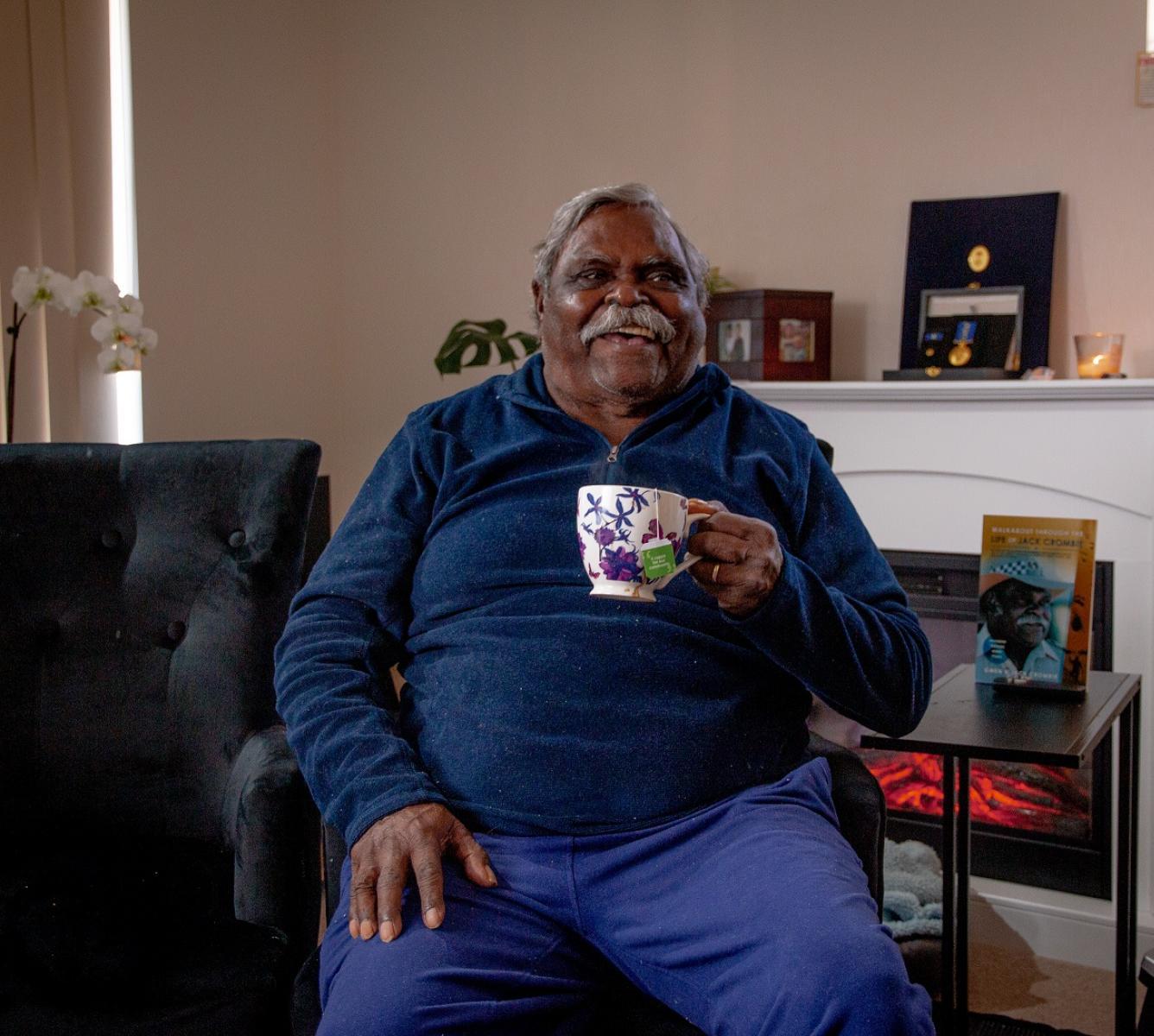 A man sitting in his living room holidng a coffee mug and smiling