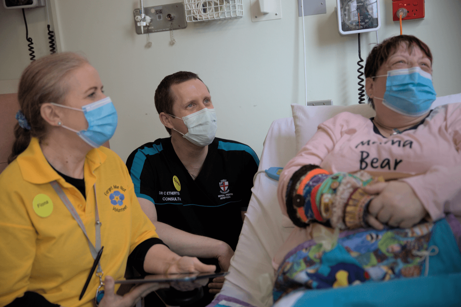 Dr Chris, a female medical staff member wearing a mask, and a patient laying in a hospital bed