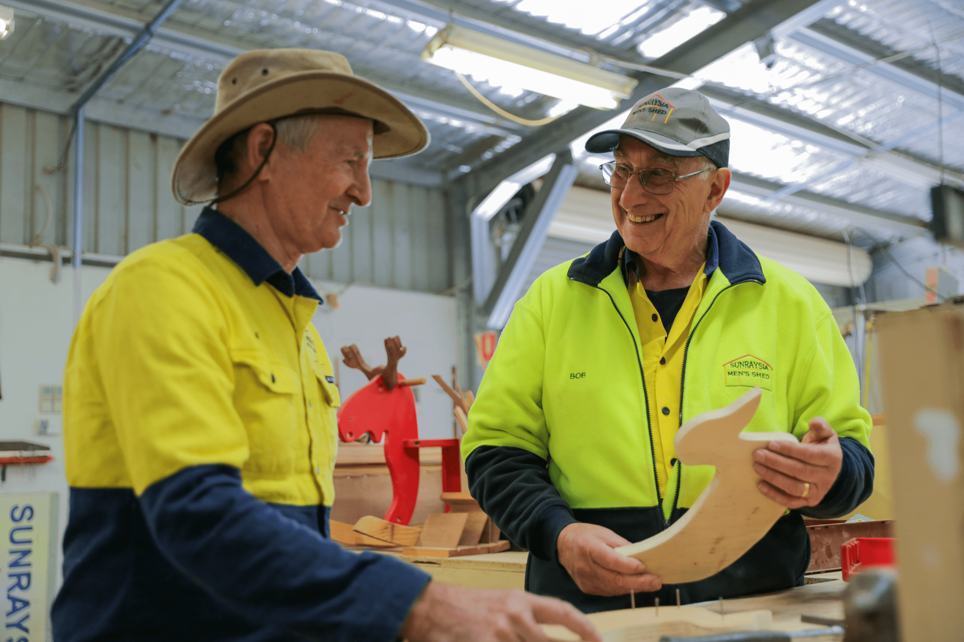 Two men in high vis smile while standing at a woodworking table at a mens shed